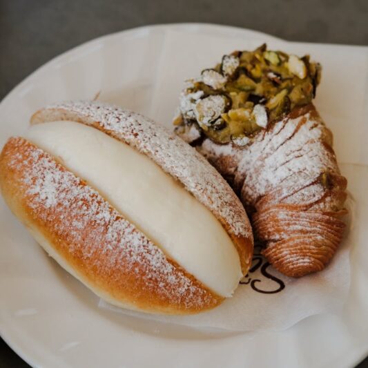 Two freshly made Italian pastries topped with powdered sugar and pistachios on a white plate.